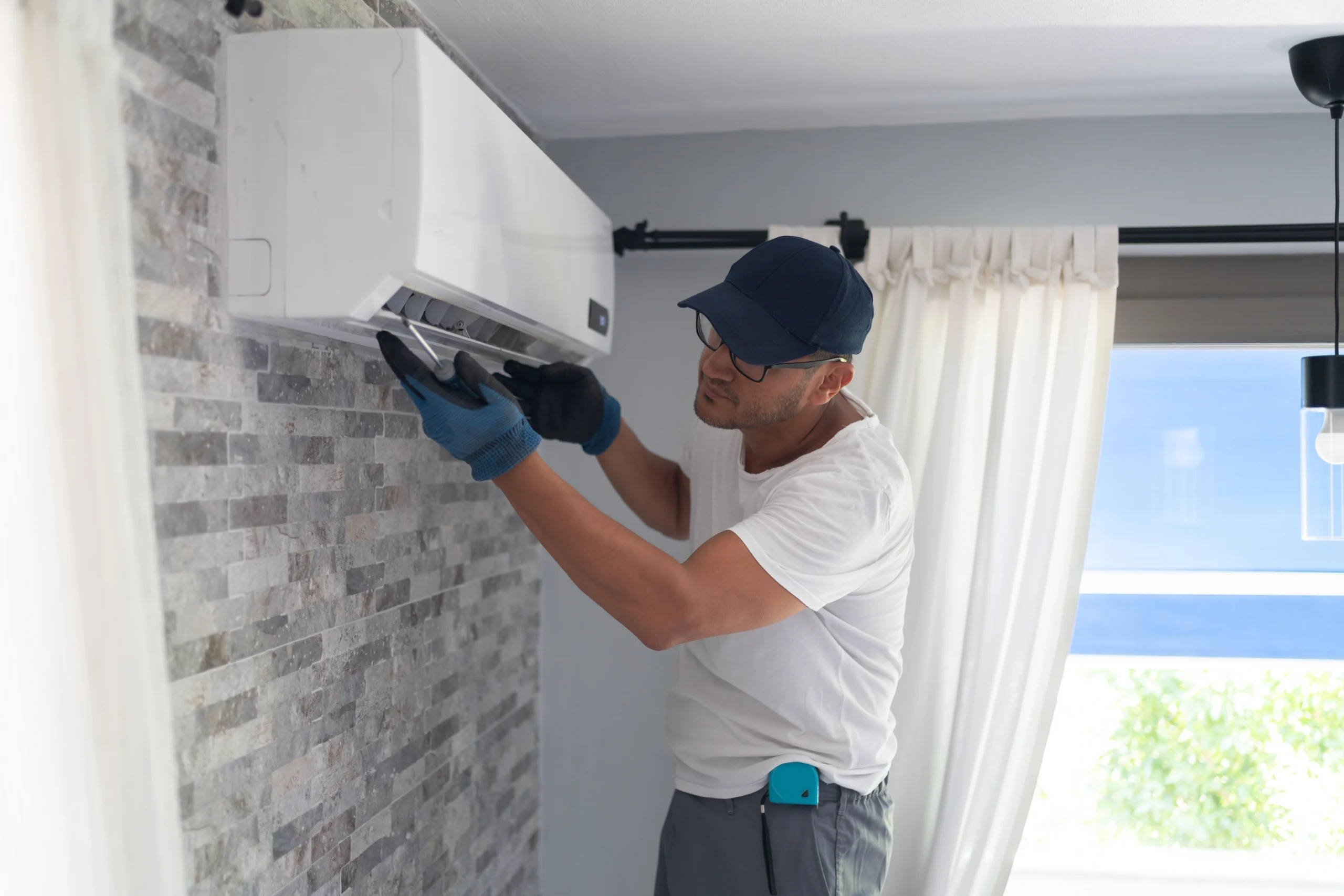 A man repairs an air conditioner in a room, focused on the unit with tools in hand.